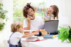 A mother sitting at a computer with her two children. She needs a garage door repair and she is trying to talk to Overhead Door Company of Huntsville. The little girl has placed a kitten on her head while her soon is sitting to her right and is playing a child's flute. She looks totally exhausted.