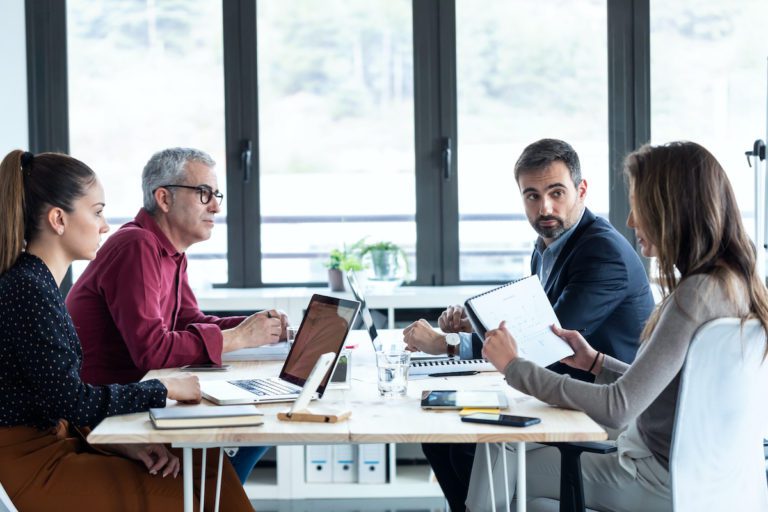 A group of architects meeting with the team from Overhead Door Company of Huntsville to discuss new garage doors to be installed. There are two men and two women seated around an rectangular table with bright light streaming in from the windows.