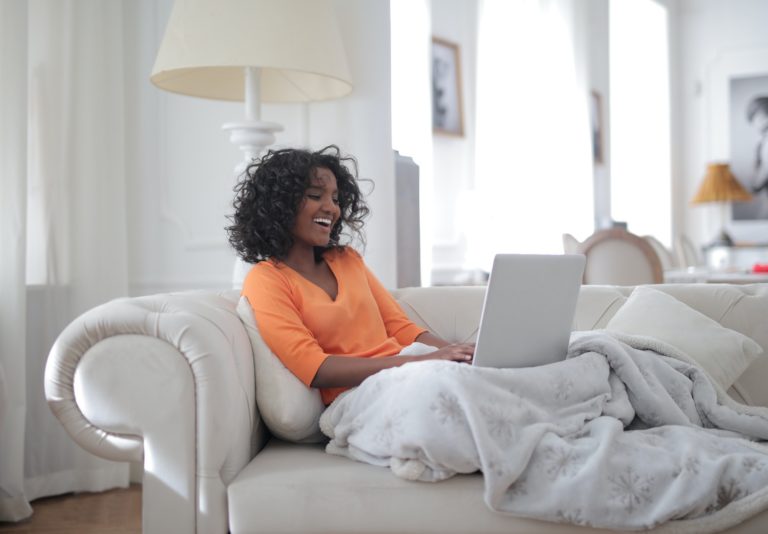 A female lady of color wearing a orange shirt sitting on a white couch happily looking at her computer for her garage door service. She is reviewing the free troubleshooting guide offered by Overhead Door Company of Huntsville/North Alabama™.