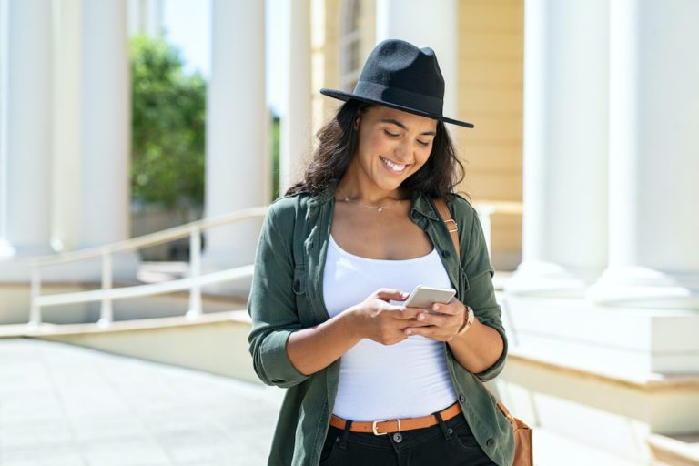 Cheerful young woman wearing black hat in city street typing a message. Hispanic trendy girl browsing internet on phone outdoor. Happy stylish girl using smartphone while travelling the city.