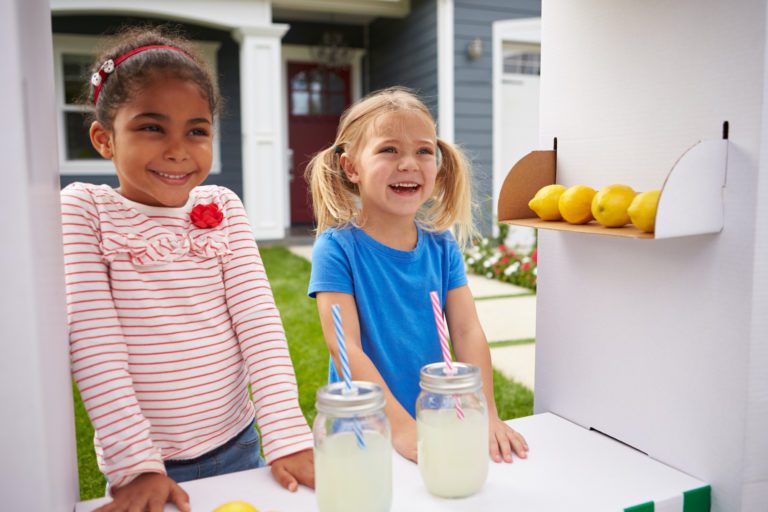 One mixed and one white colored girl smiling happily as they are outside their home with a lemonade stand. A beautiful summer day. Two glasses of lemonade sitting in front of them. There home just had a white residential garage door installed by Overhead Door Company of Huntsville/North Alabama. The number one garage door repair and installation company that services Huntsville, Madison, Athens and all of Limestone and Madison Counties.