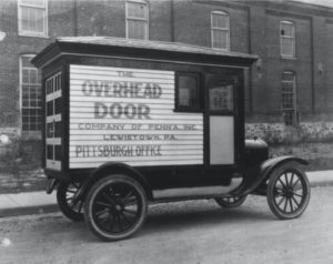 Old Vintage Car carrying an Overhead Door Company Sample. On the side of the old car the words Overhead Door appear. This is about the history of the first garage door company and how they made their sales around the world.