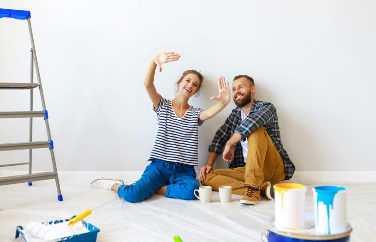 Lady with her hands up visualizing a new garage door as her husband smiles next to her. They are in the middle of a home remodel and are sitting in a white room with paint cans.