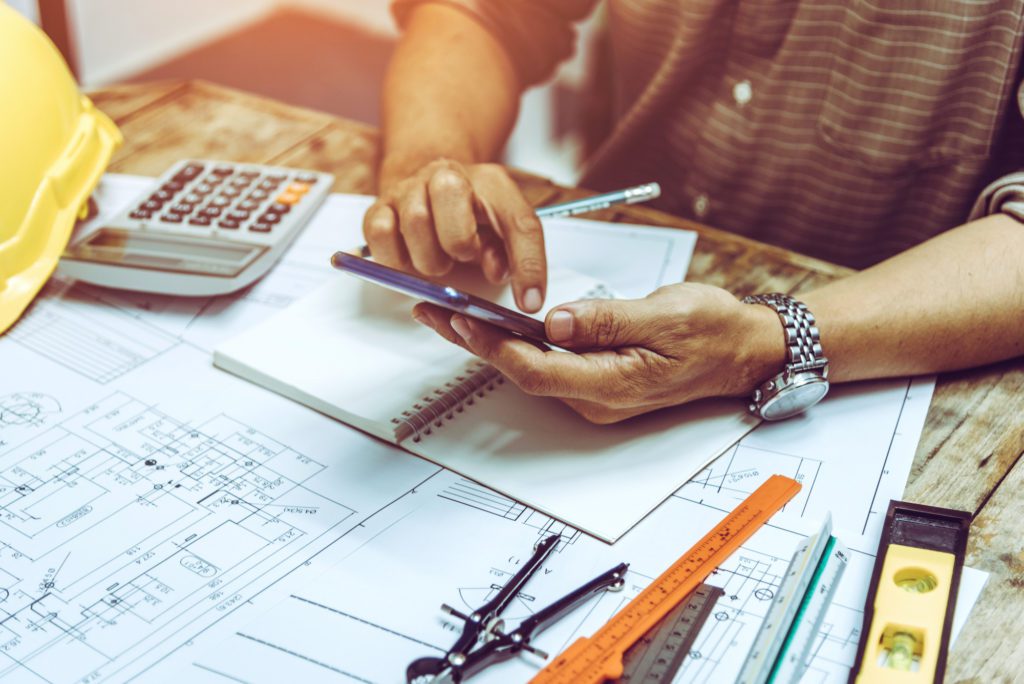 Man calculating the measurements on his garage door before deciding which new garage door he wants installed from Overhead Door. He is a white male with a calculator and a messy desk with house plans, pencils and rulers scattered everywhere.