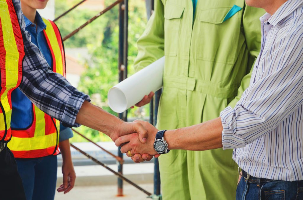 A garage door service technician wearing a safety vest shaking hands with an architect and lead man on a project job location.
