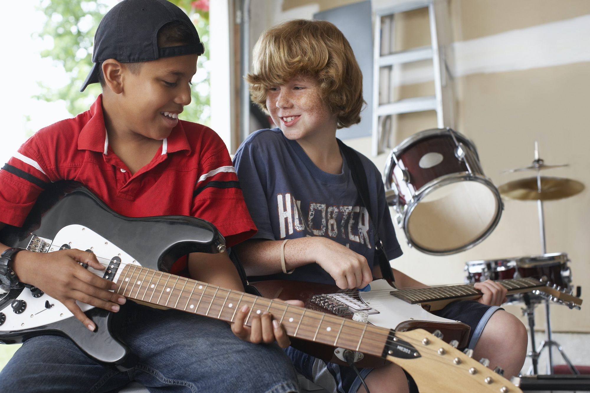 Two kids playing the guitar in a garage. One child is of color the other white. The children are happy and smiling. Their age is 10.