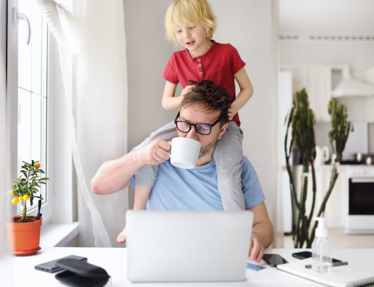 Adorable setting. A child with a bright red shirt sitting on top of his father's shoulders. The father is on the computer looking at garage door pictures while drinking a cup of coffee. The male child is looking down at his fathers computer and smiling. It appears to be early in the morning.