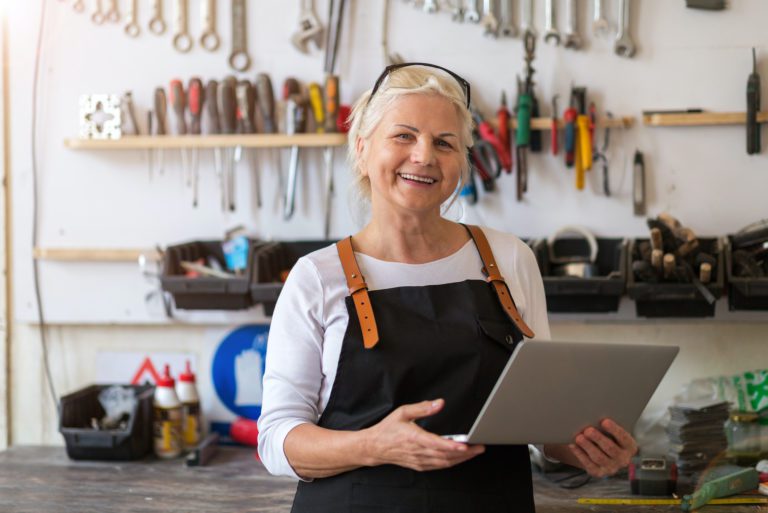 Lady in her sixties standing in her garage with her shop apron on. She is looking at her computer and smiling. Behind her is a peg board full of her tools. She is online shopping for garage door products from Overhead Door.